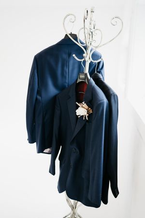 Navy blue toddler suit jacket with boutonniere displayed on white decorative hanger against white background.