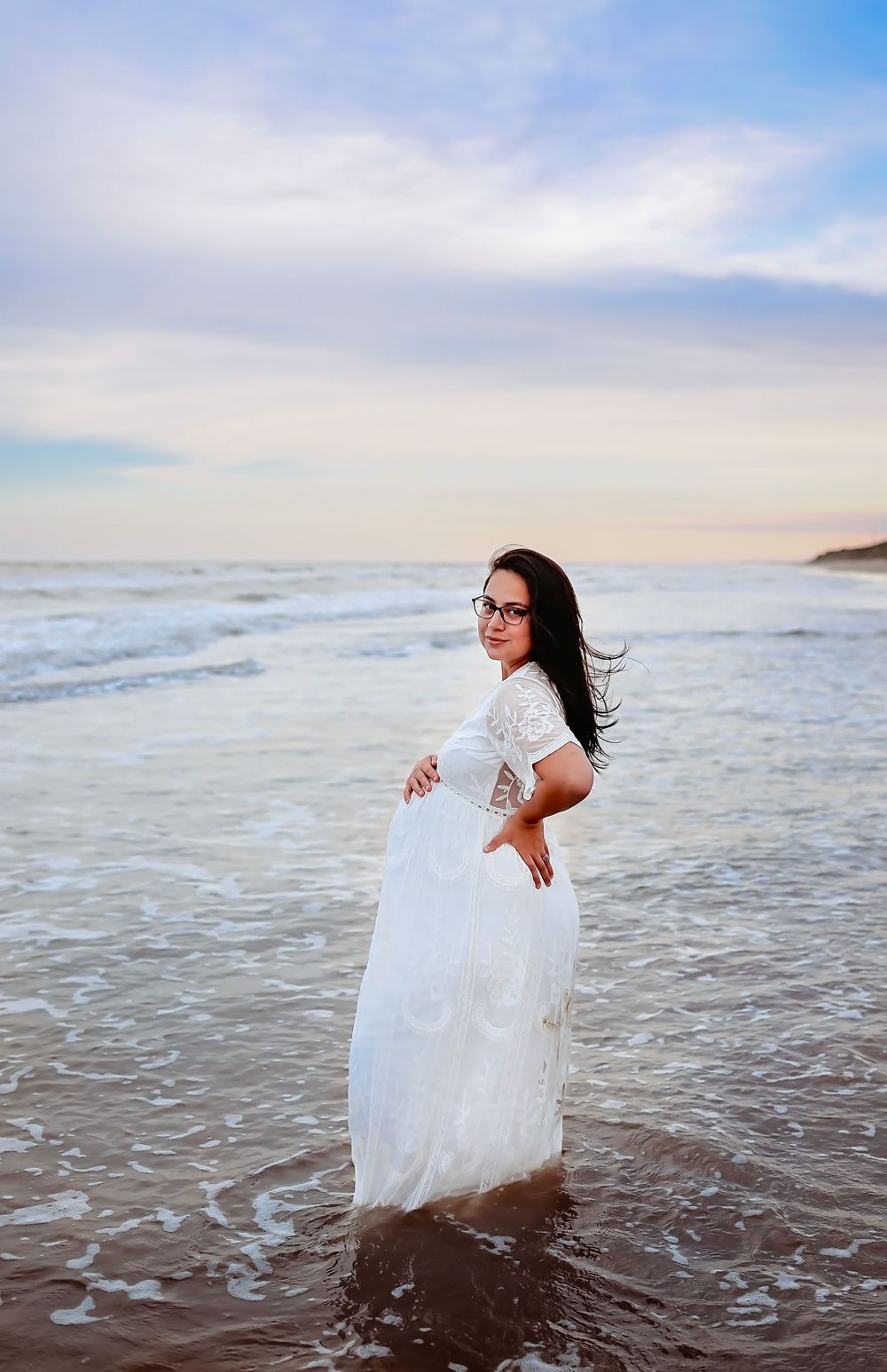 pregnant mama on beach holding belly in white dress during maternity session