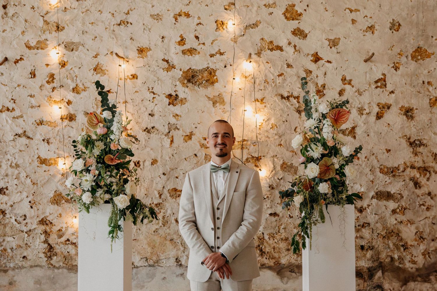 Smiling groom in a beige suit with bow tie standing between two floral arrangements against a golden textured wall.
