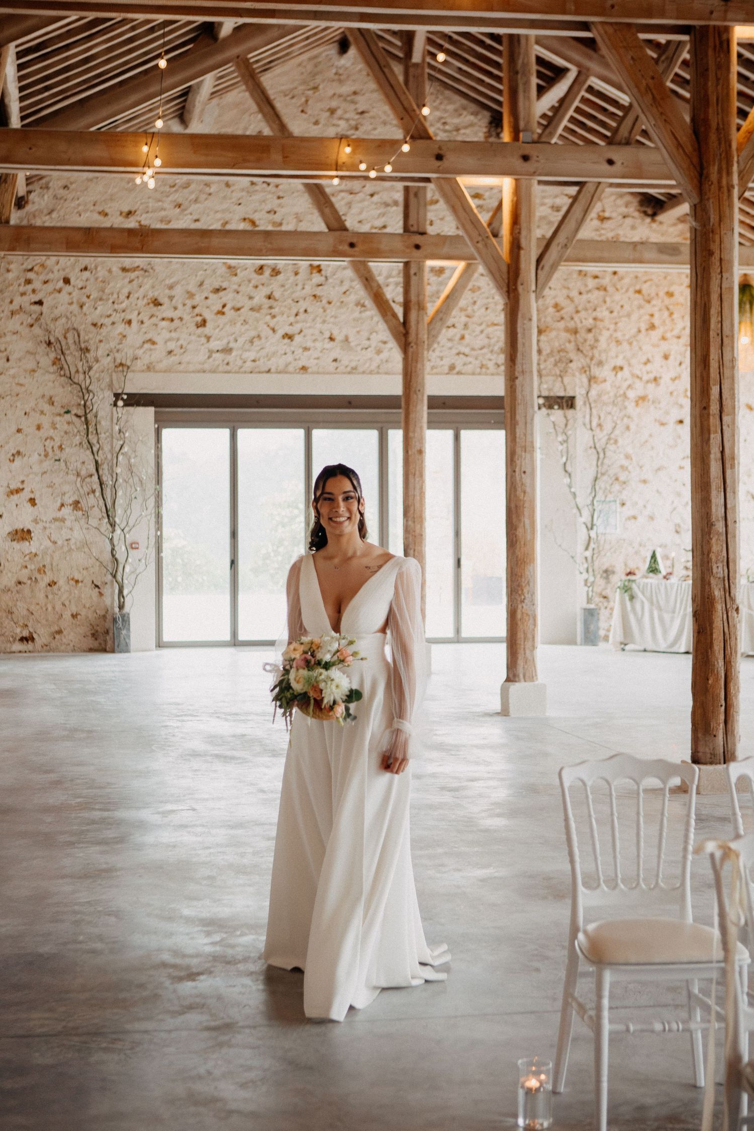 Elegant bride in a long-sleeve white wedding dress holding a bouquet in a rustic barn venue with string lights.