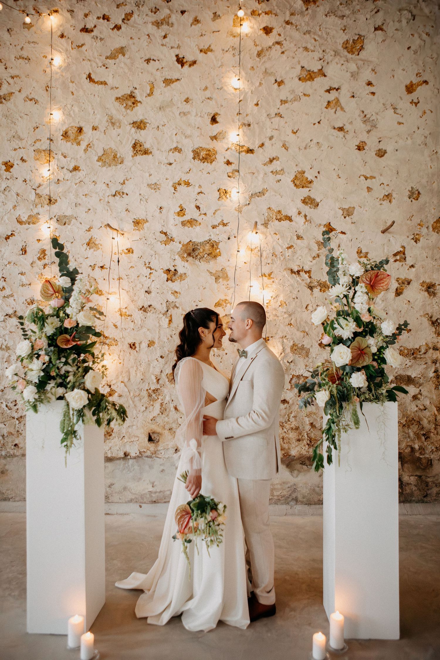 Bride and groom standing together between floral pedestals and candles against a warmly lit textured stone wall.