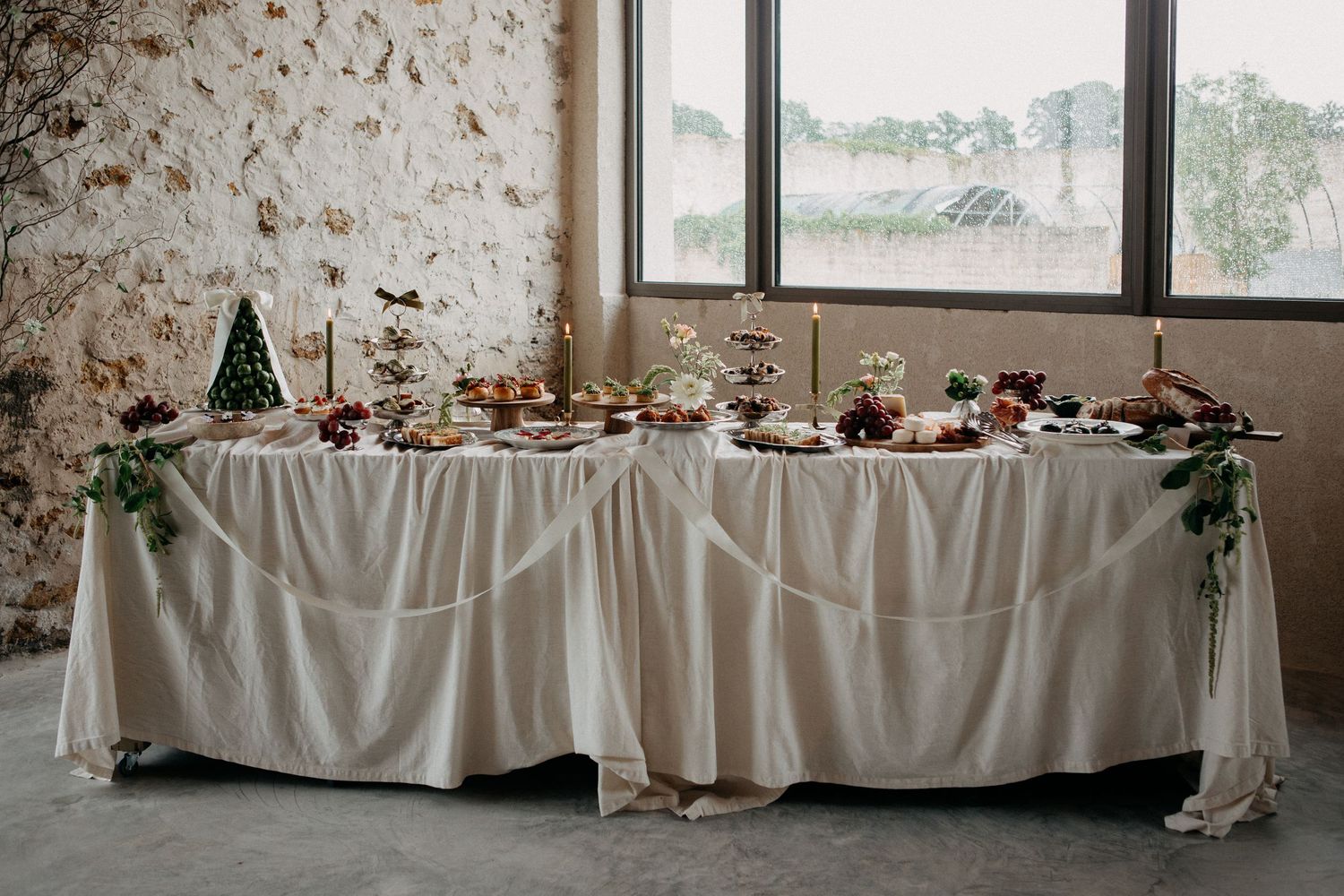 Elegant wedding reception grazing table with white draping, floral garlands, and candles near a large window.