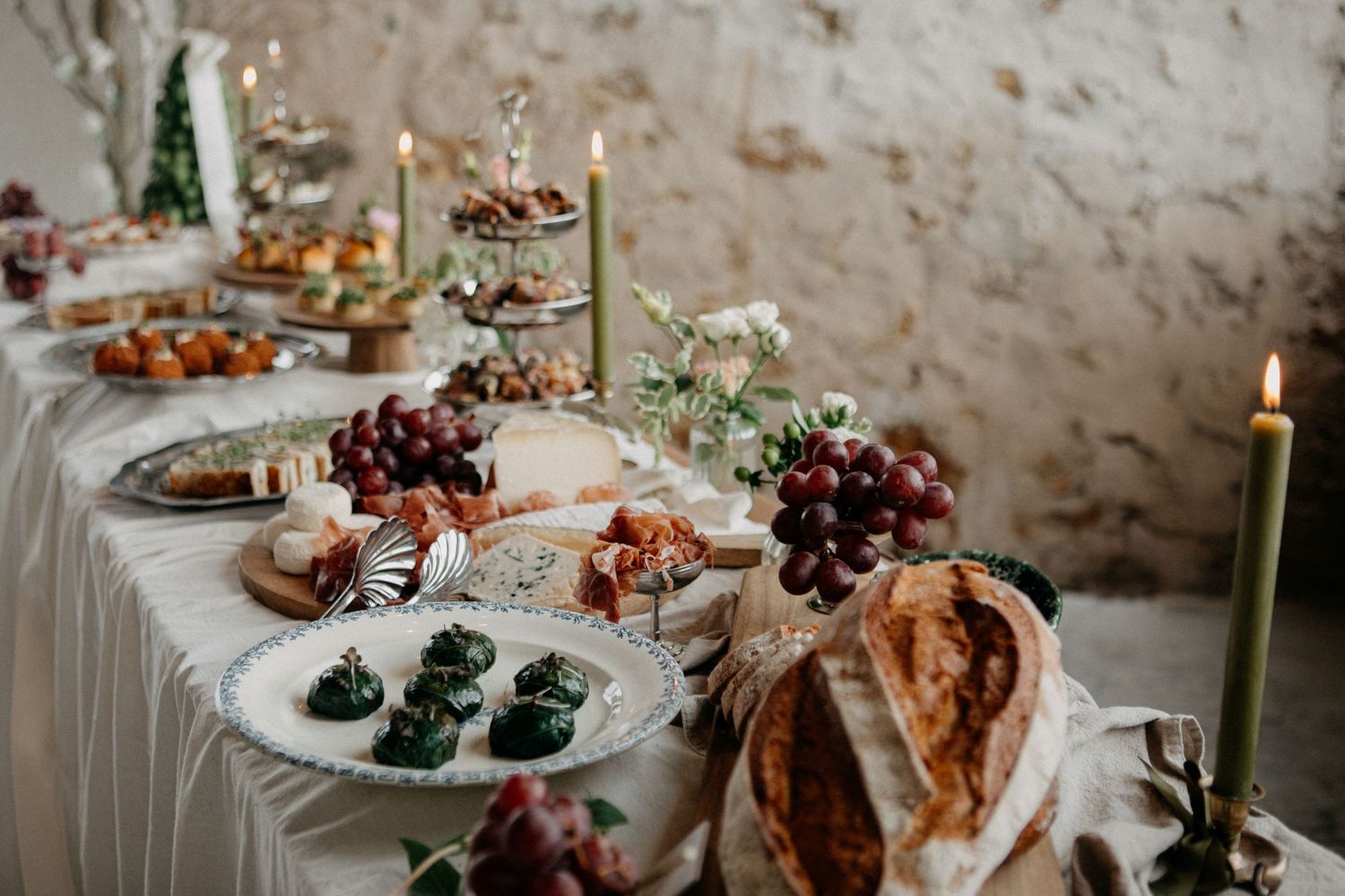 Beautifully styled wedding grazing table featuring fruits, bread, candles, and greenery against a rustic stone wall.