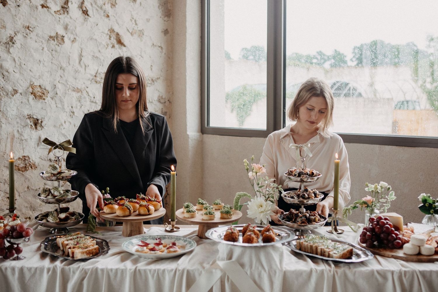Two women arranging a wedding dessert table with tiered stands, pastries, and elegant decorations in a rustic venue.