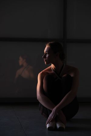 Contemplative dancer in black leotard sitting in low light with reflection on wall.