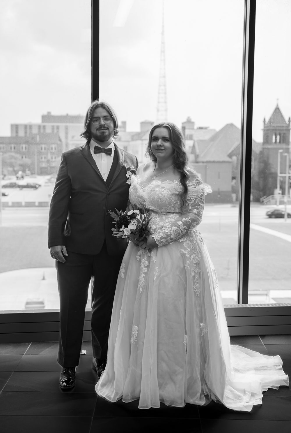 A black and white wedding portrait featuring a couple against a window backdrop with city views.