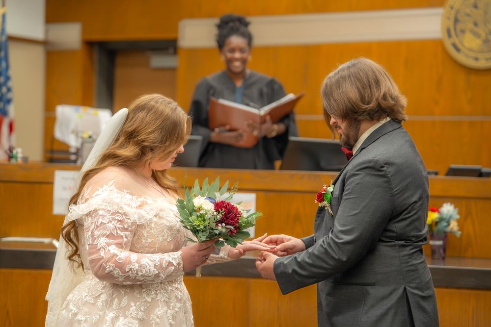 A bride in a lace dress and groom in suit sign marriage documents in a courthouse setting.