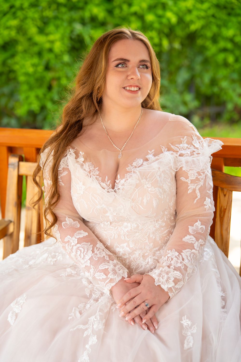 A bride in a white lace wedding dress poses on a wooden bench in a garden setting.