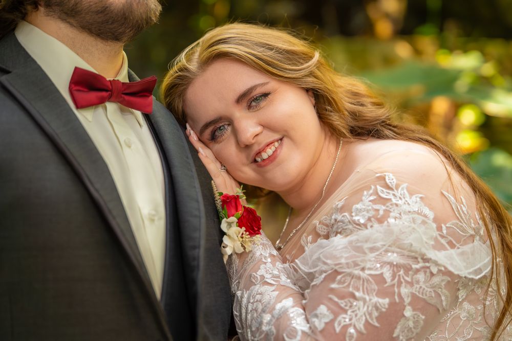 A bride in a white lace dress smiles warmly while standing next to her groom wearing a dark suit with red bowtie.