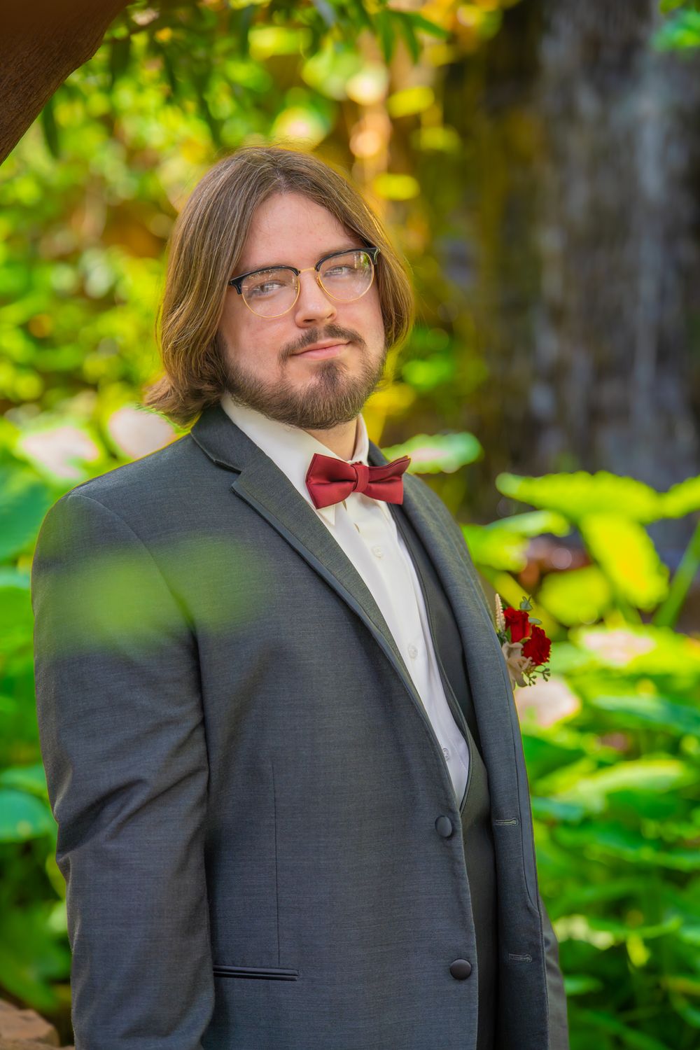 Wedding guest wearing dark suit with red bowtie stands in lush garden setting.