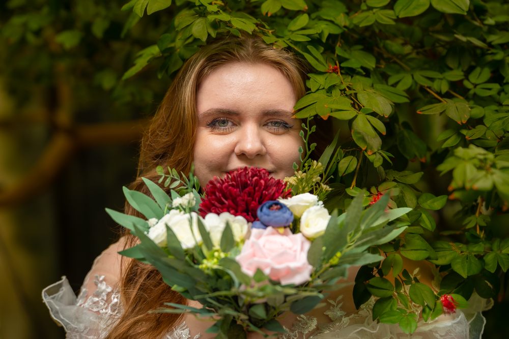 Smiling figure holding vibrant bouquet of red, white and pink flowers against leafy backdrop.