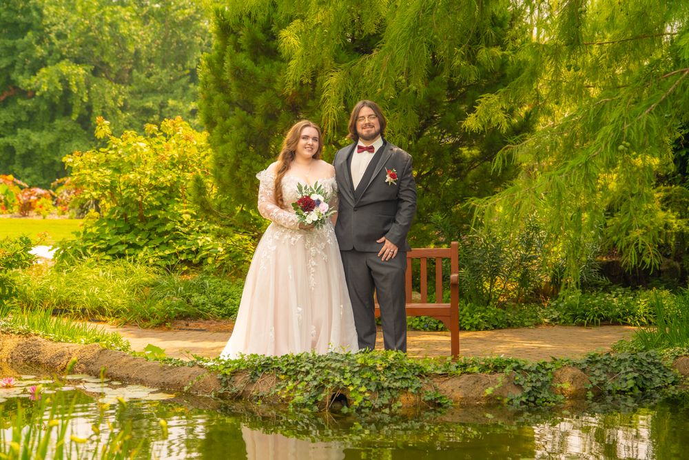 Newlyweds pose together on garden path beside reflective pond with evergreen trees in background.
