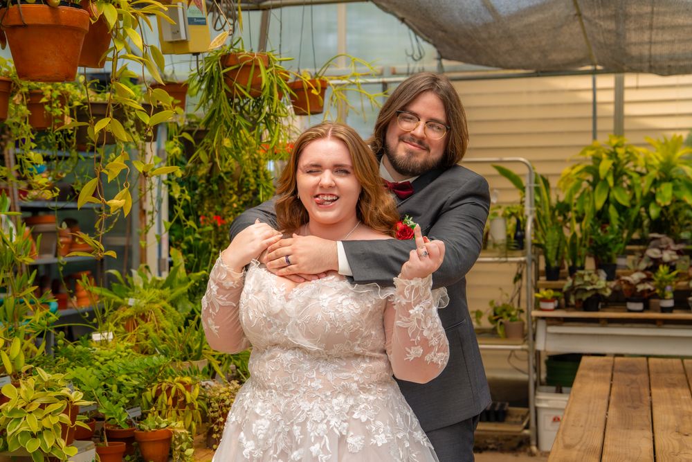 A laughing couple posing together in a greenhouse filled with lush plants and natural sunlight.