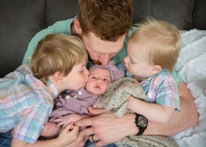 Three small children wearing pastel colors snuggle together while lying on a soft grey surface.