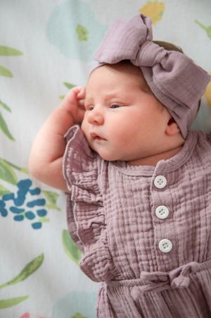 Baby in gray textured outfit with white buttons lies against floral background while wearing matching bow headband.
