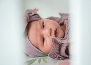 Sweet baby wearing pink lies in crib looking at camera while resting on pastel bedding.
