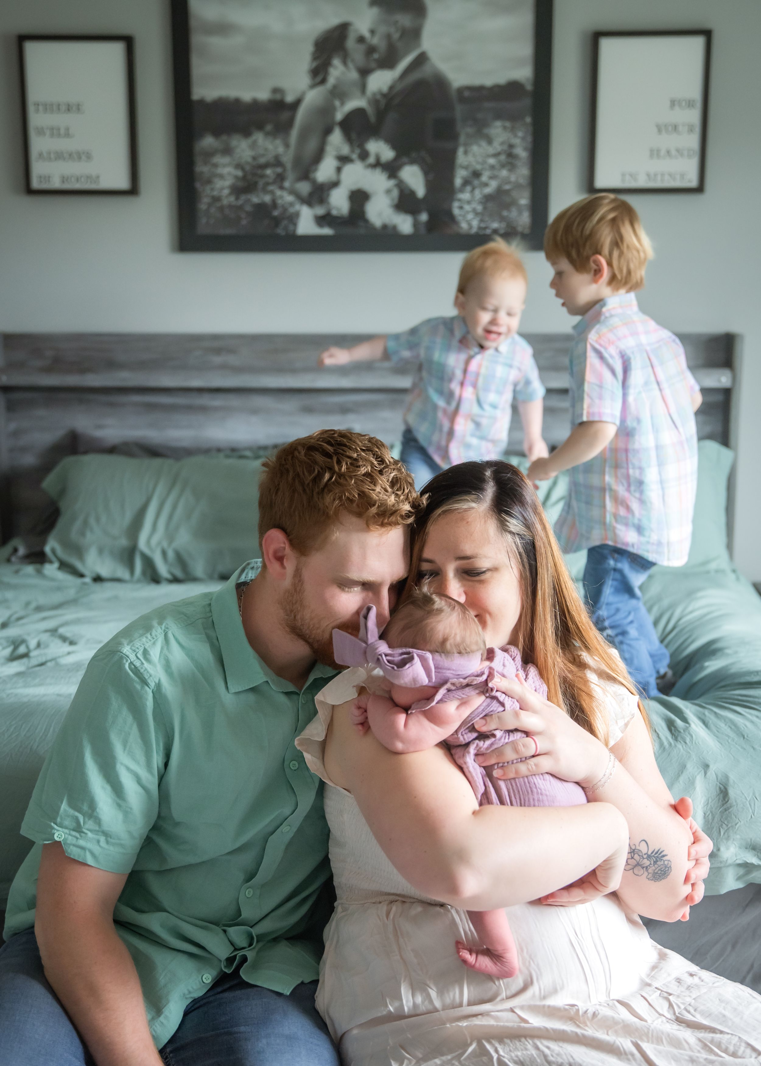 Sweet candid family moment on a bed with two children playing in background and a framed wedding photo on the wall.