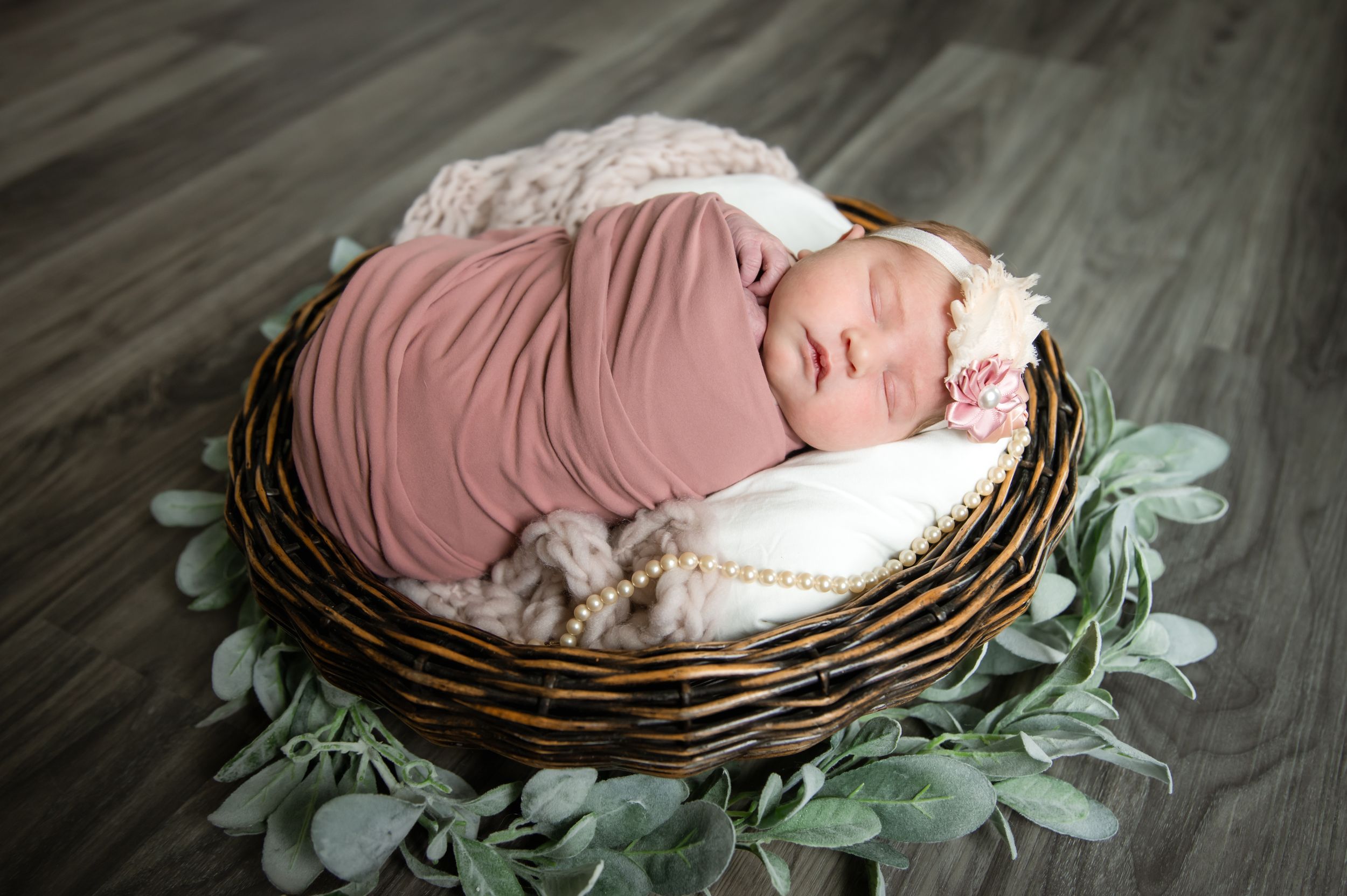 Newborn baby swaddled in pink fabric sleeps peacefully in a woven basket nest surrounded by eucalyptus leaves.