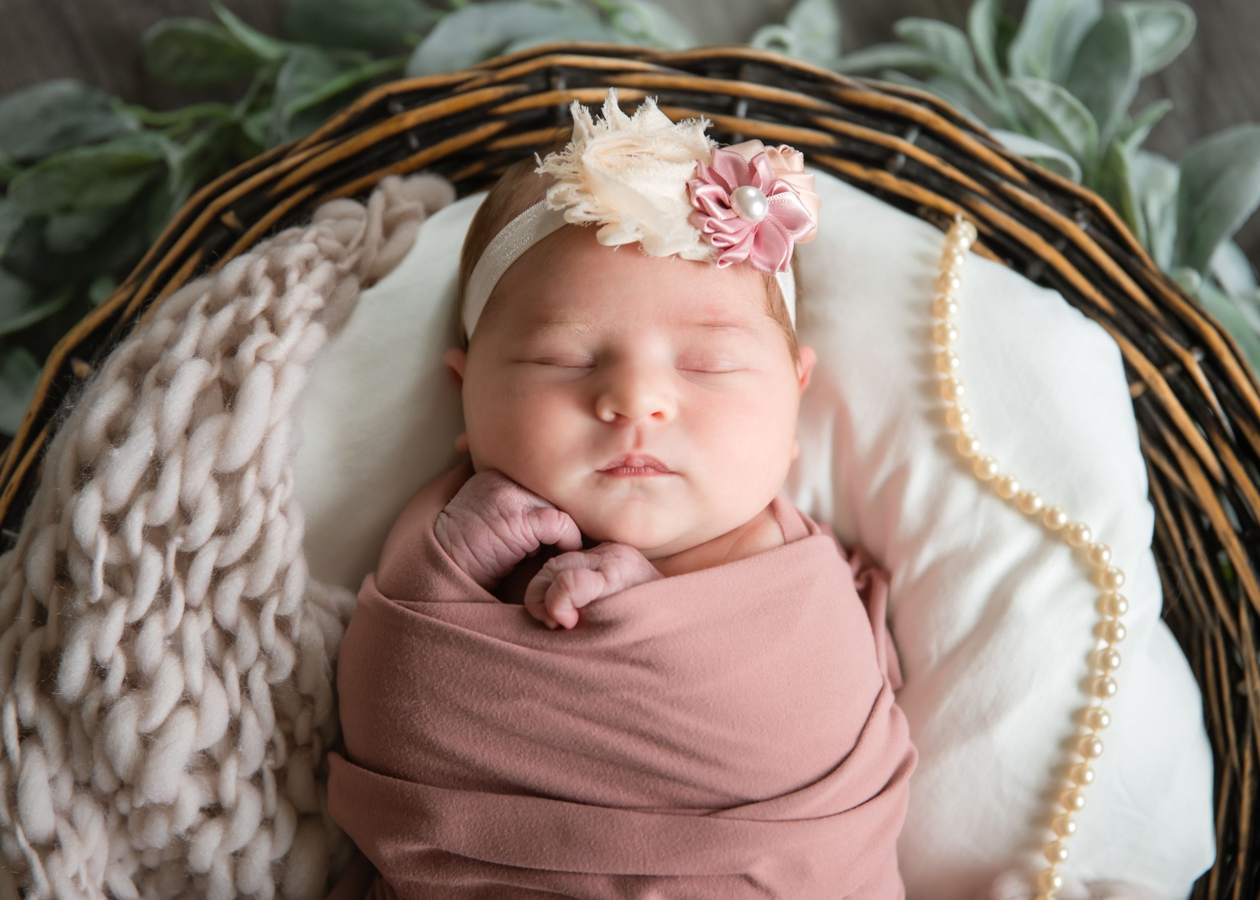 Sleeping newborn baby wrapped in pink swaddle wearing floral headband in wicker basket with pearls.