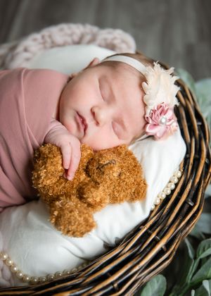 Sleeping baby in pink outfit cuddles with brown teddy bear in wicker basket lined with white cushioning.