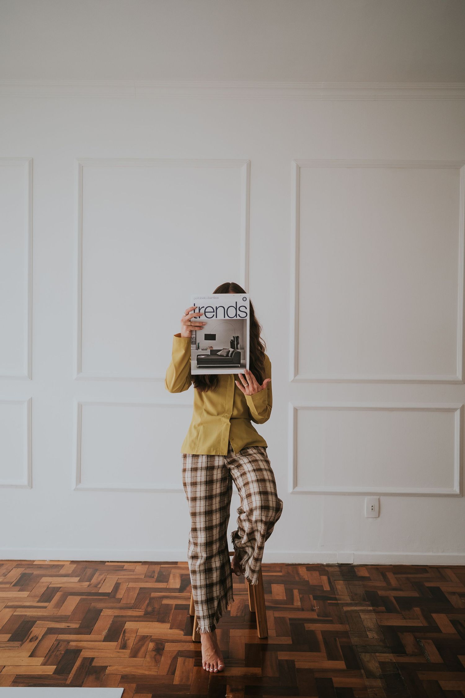 Person in yellow top and plaid pants reading magazine on herringbone wood floor.