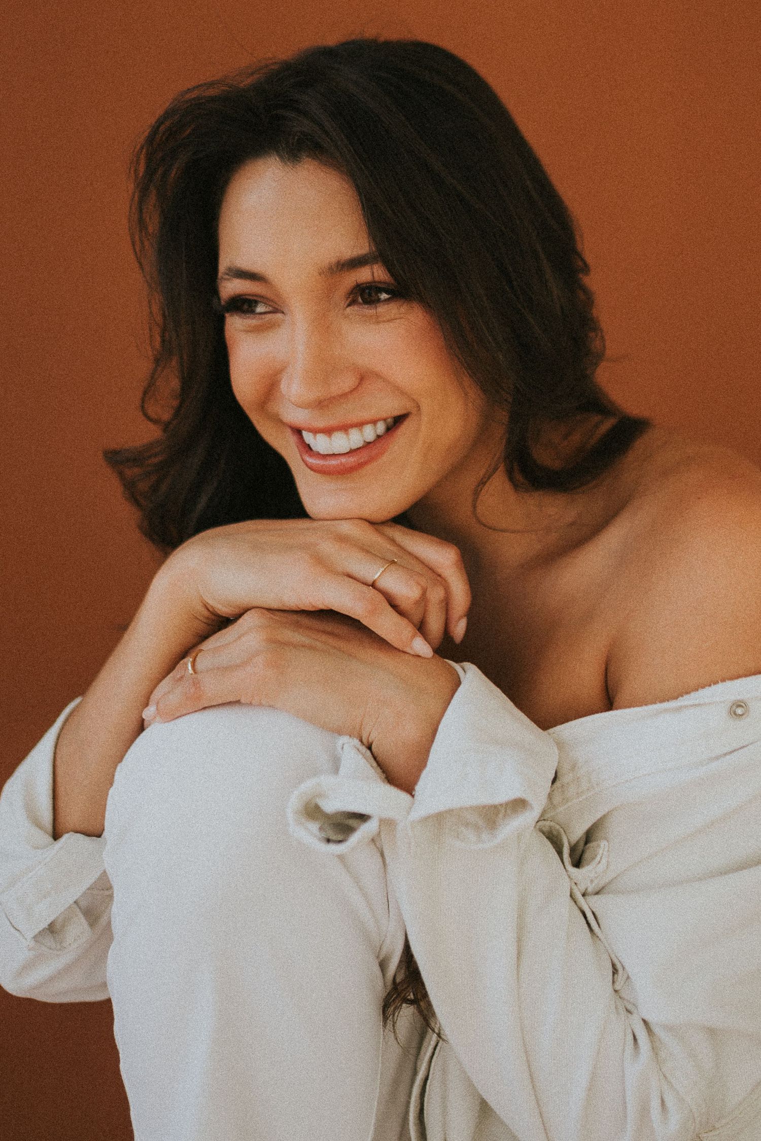 Intimate portrait against warm brown background wearing white off-shoulder top.