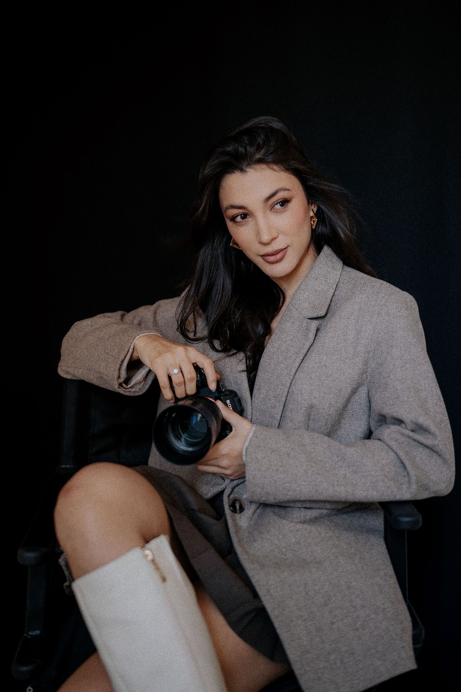 Professional photographer in grey blazer holding camera against dark backdrop.