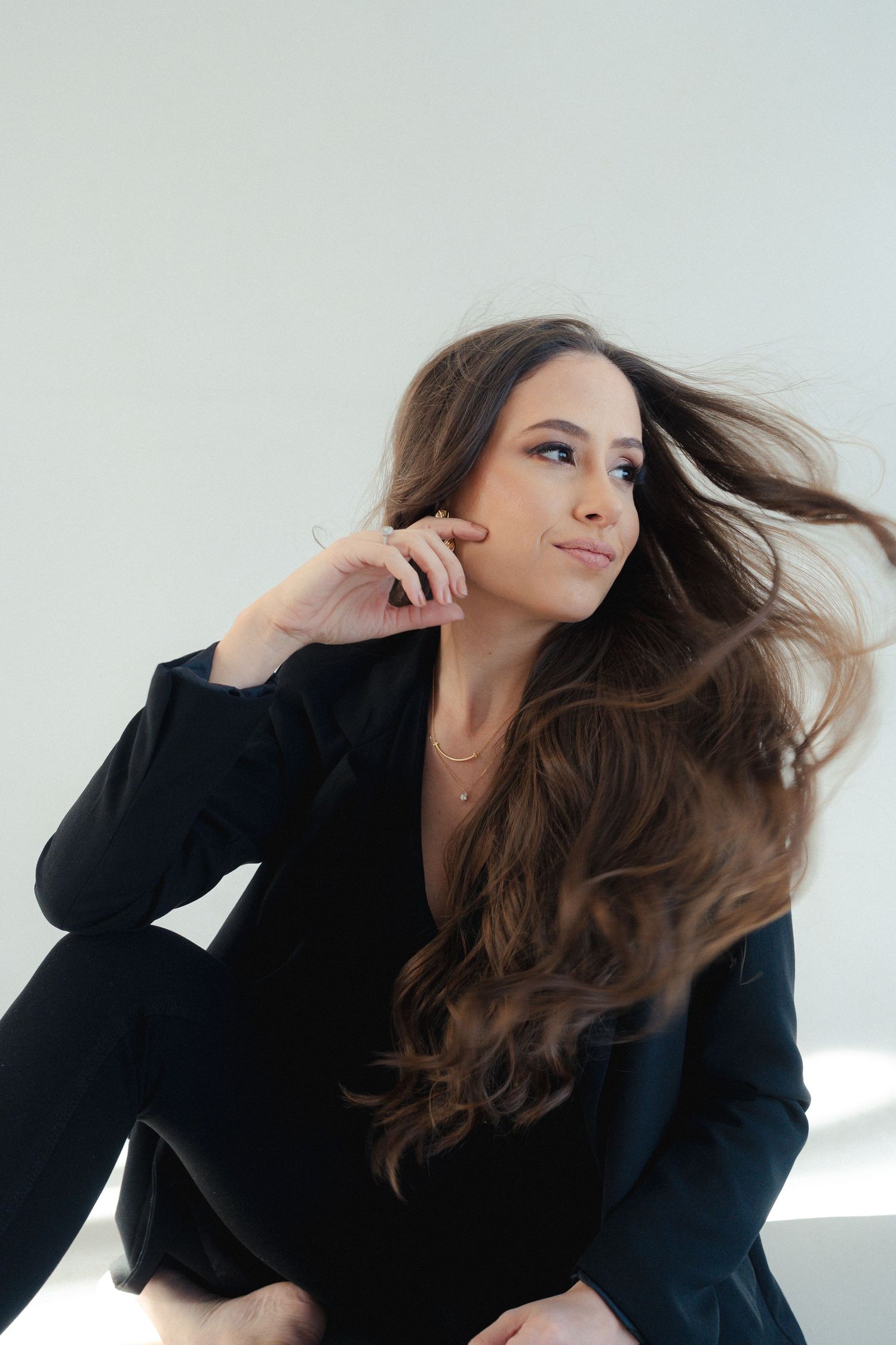 Professional portrait of a model in a black blazer with flowing long brown hair against a light background.