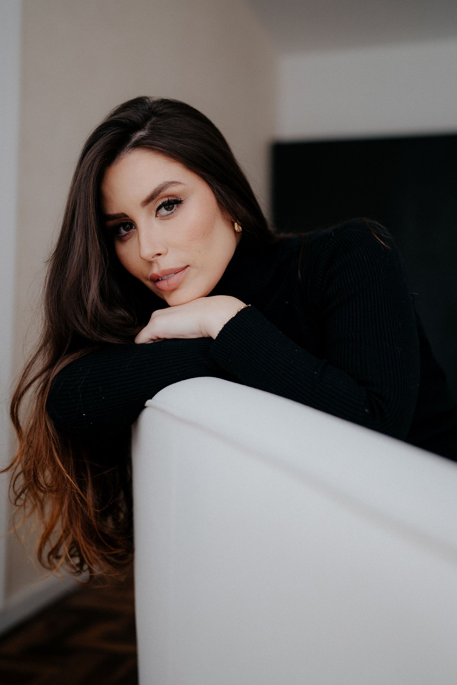 Professional portrait featuring dark long hair against white couch in minimalist setting.