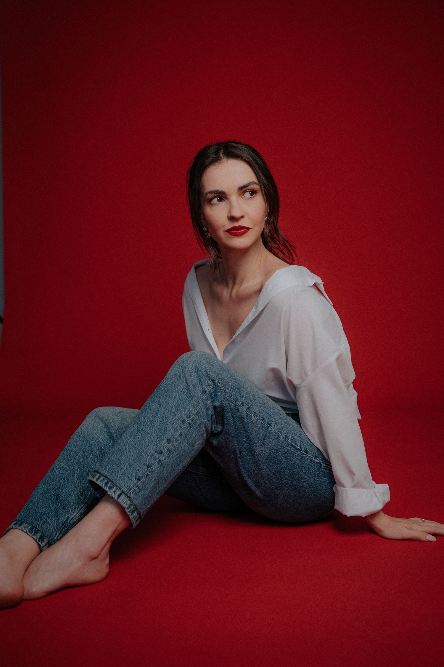 Fashion portrait of a model in casual denim and white shirt seated on red backdrop.