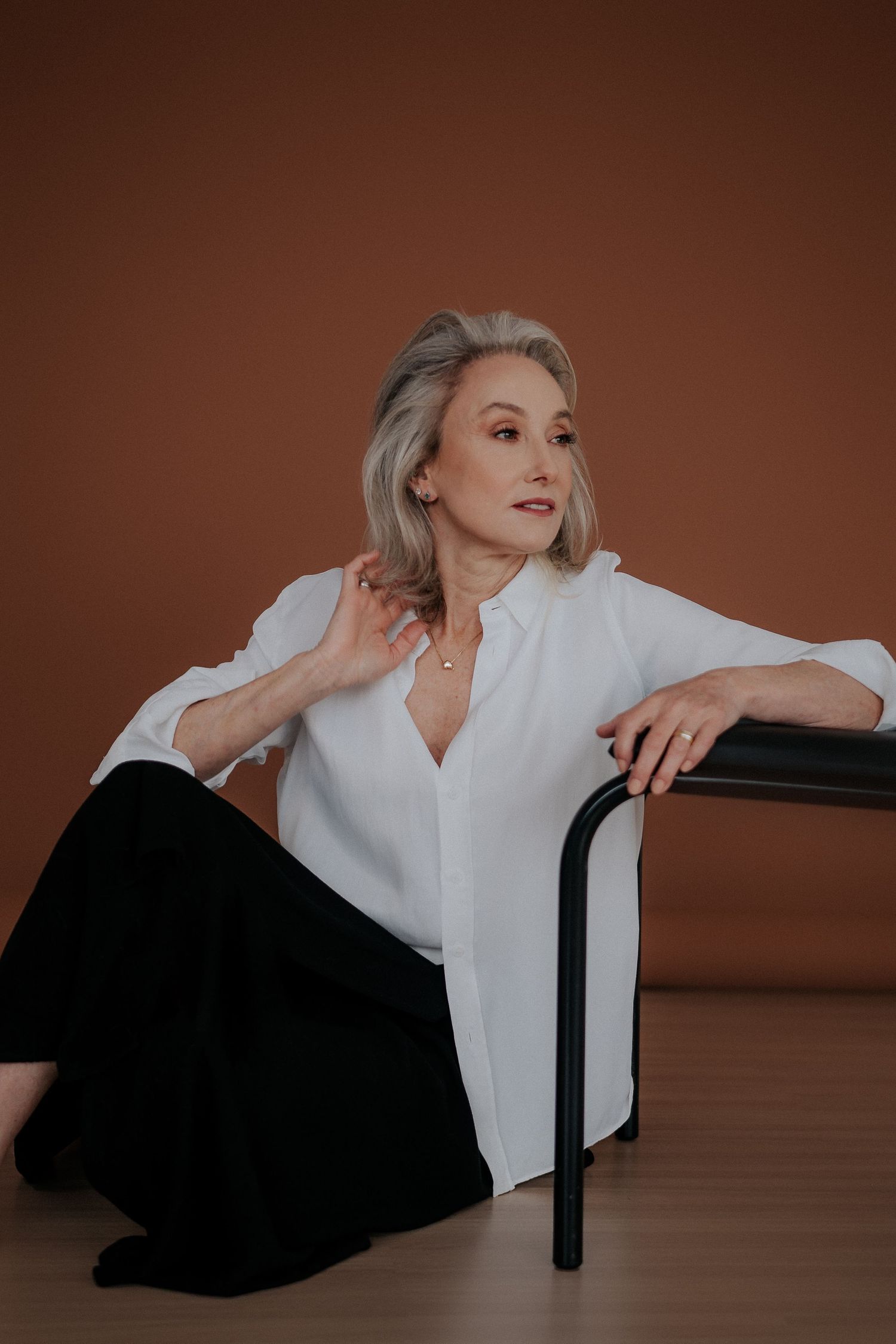 Professional studio portrait of woman in white blouse and black pants posed by chair.
