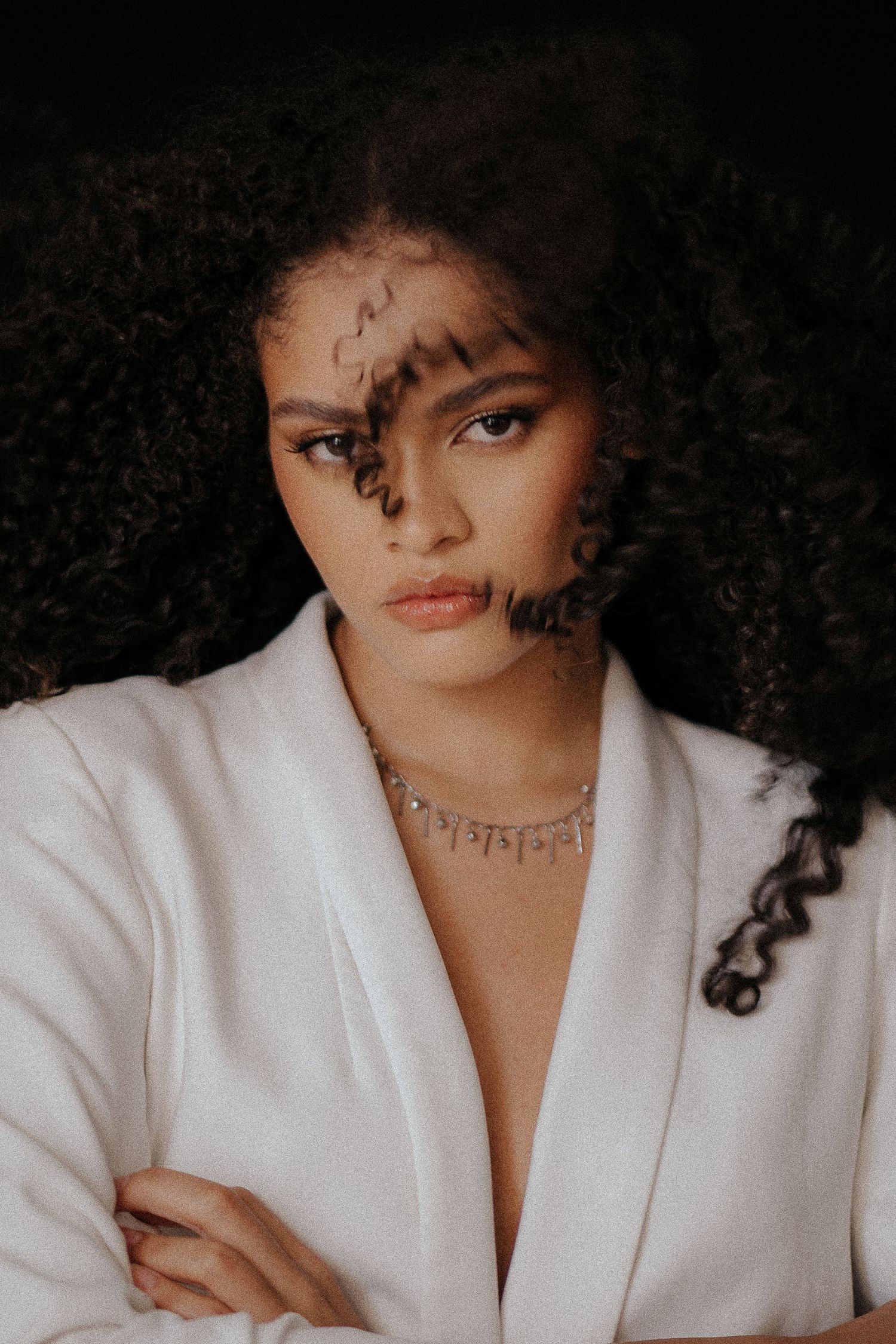 Dramatic portrait in white blazer with natural curly hair against dark background.