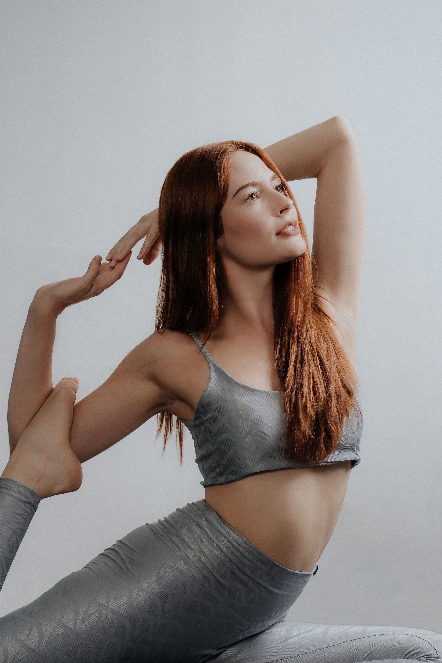 Athletic woman in grey workout attire stretching in a yoga pose against white backdrop.
