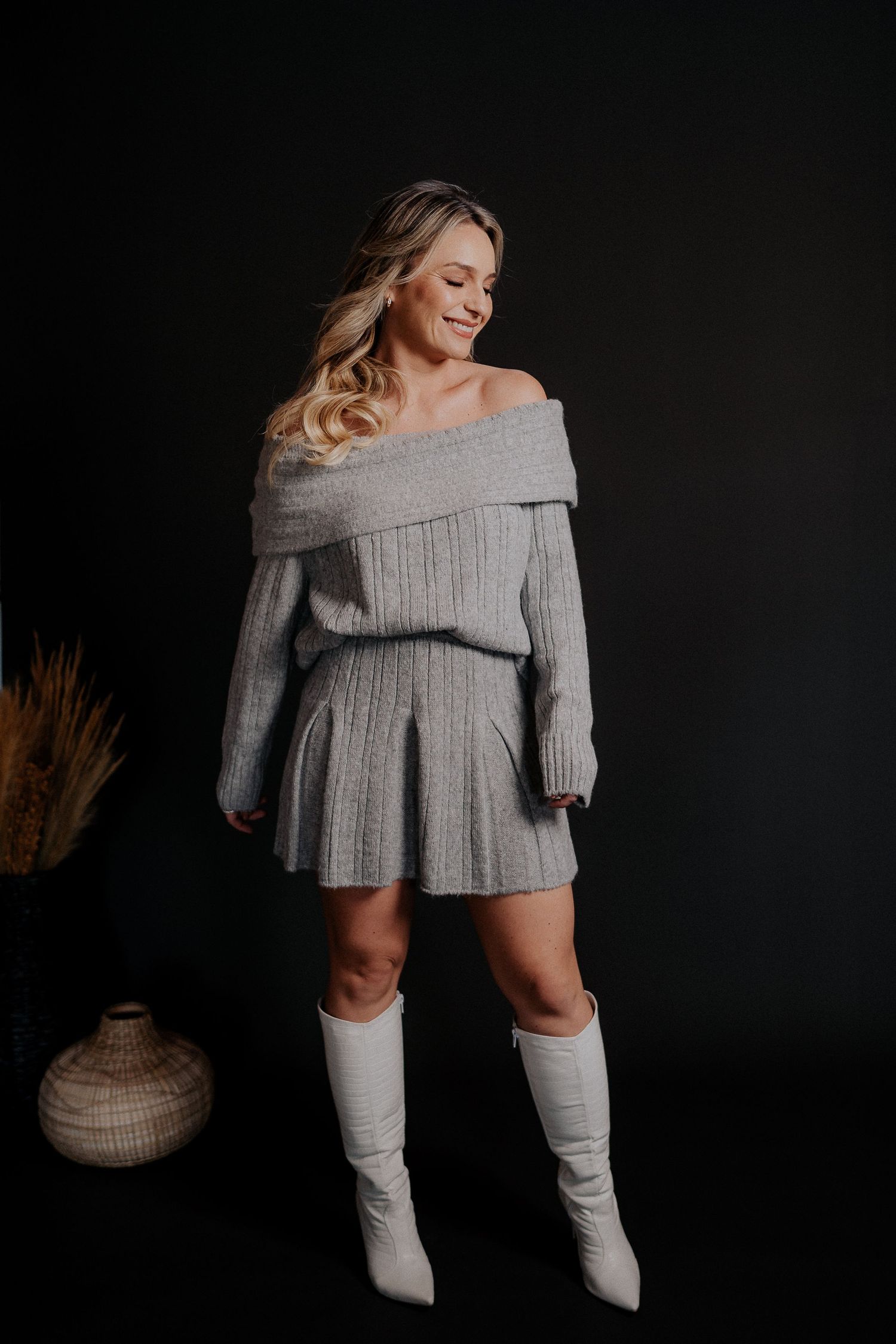 Fashion model wearing grey sweater dress and white boots in moody studio setting.
