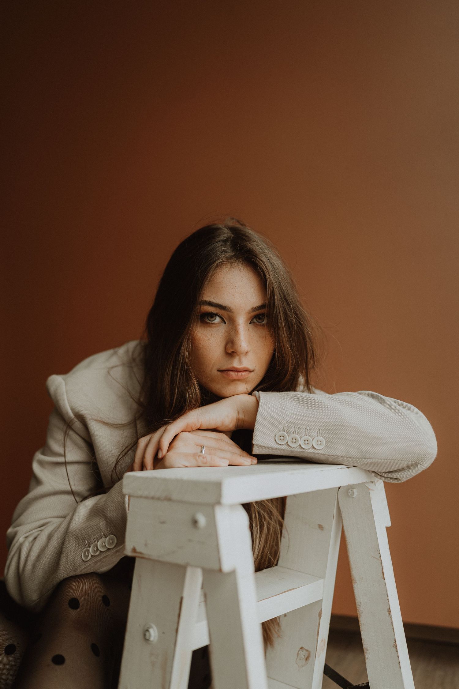 Minimalist portrait series with subject resting on white wooden stool against warm backdrop.