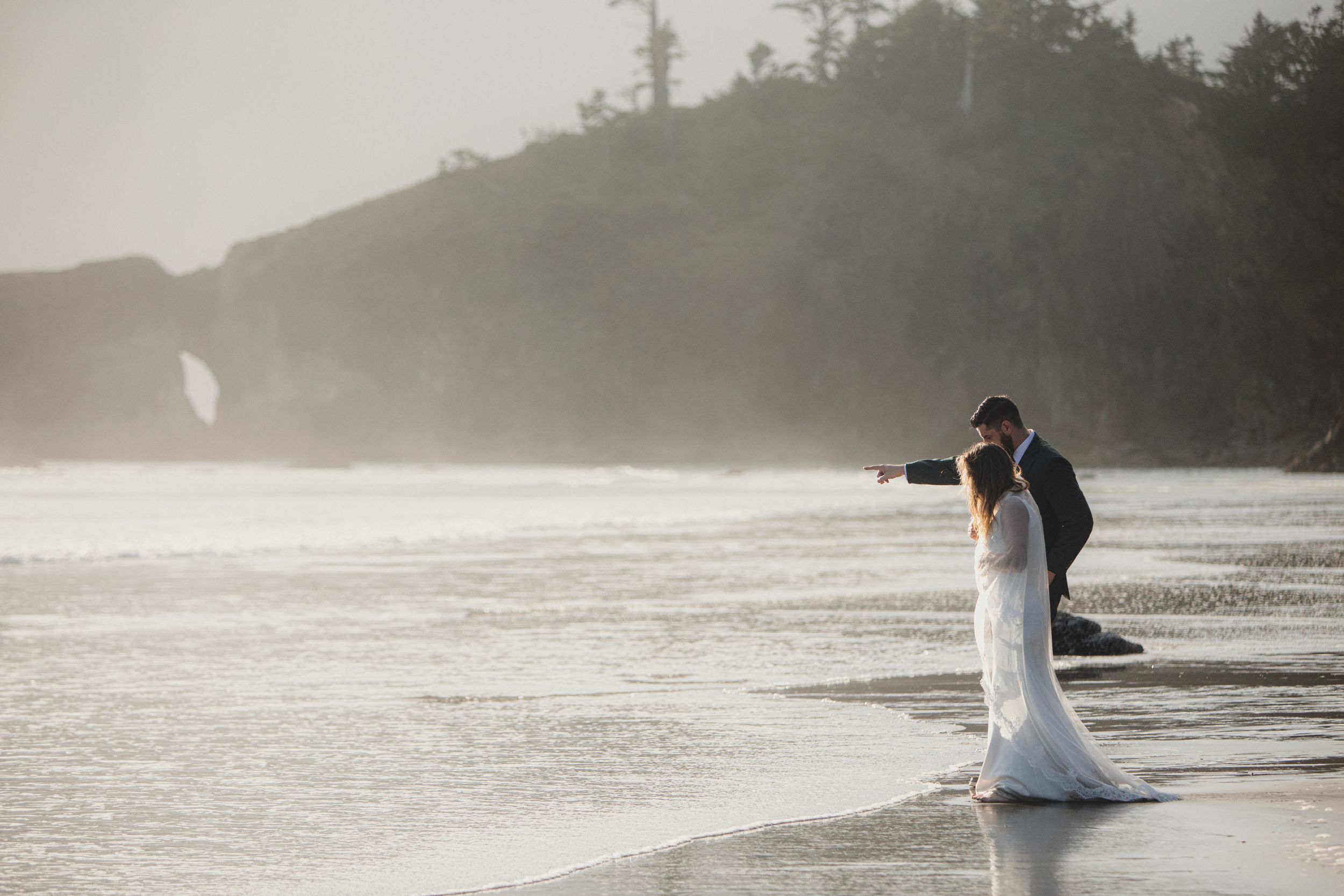 A couple in wedding attire embraces on a misty beach at sunset with cliffs in the background.