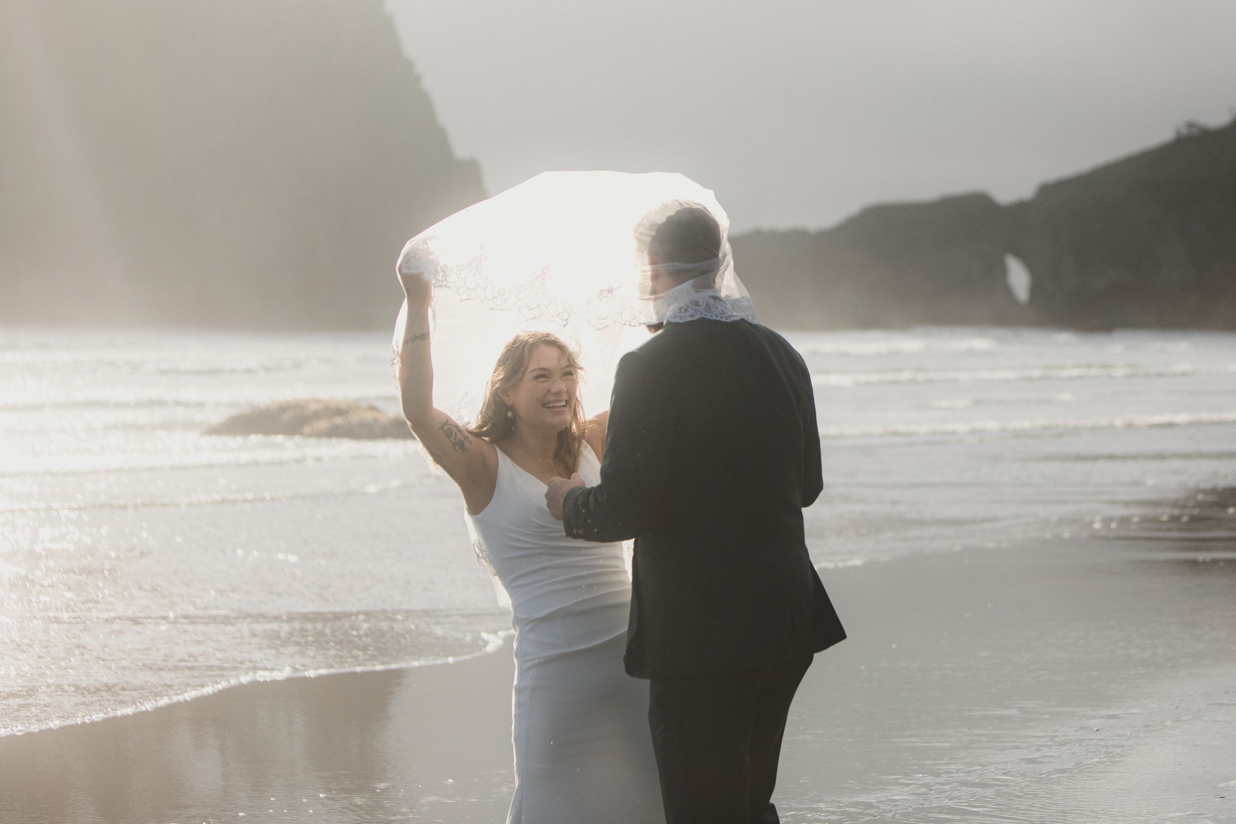 Couple walking on misty beach at sunset holding hands with ocean waves rolling onto shore in background.