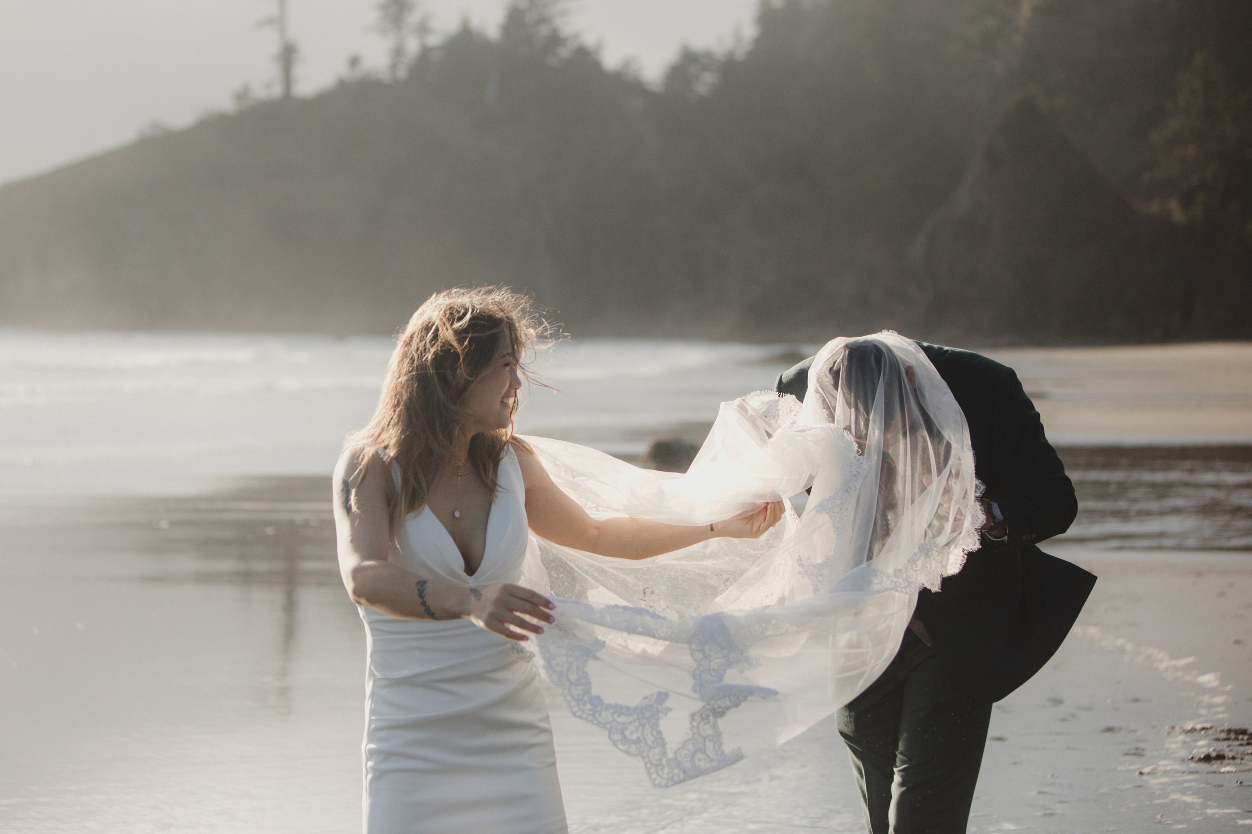 A couple in wedding attire shares a romantic moment on a misty beach with soft sunlight creating a dreamy atmosphere.