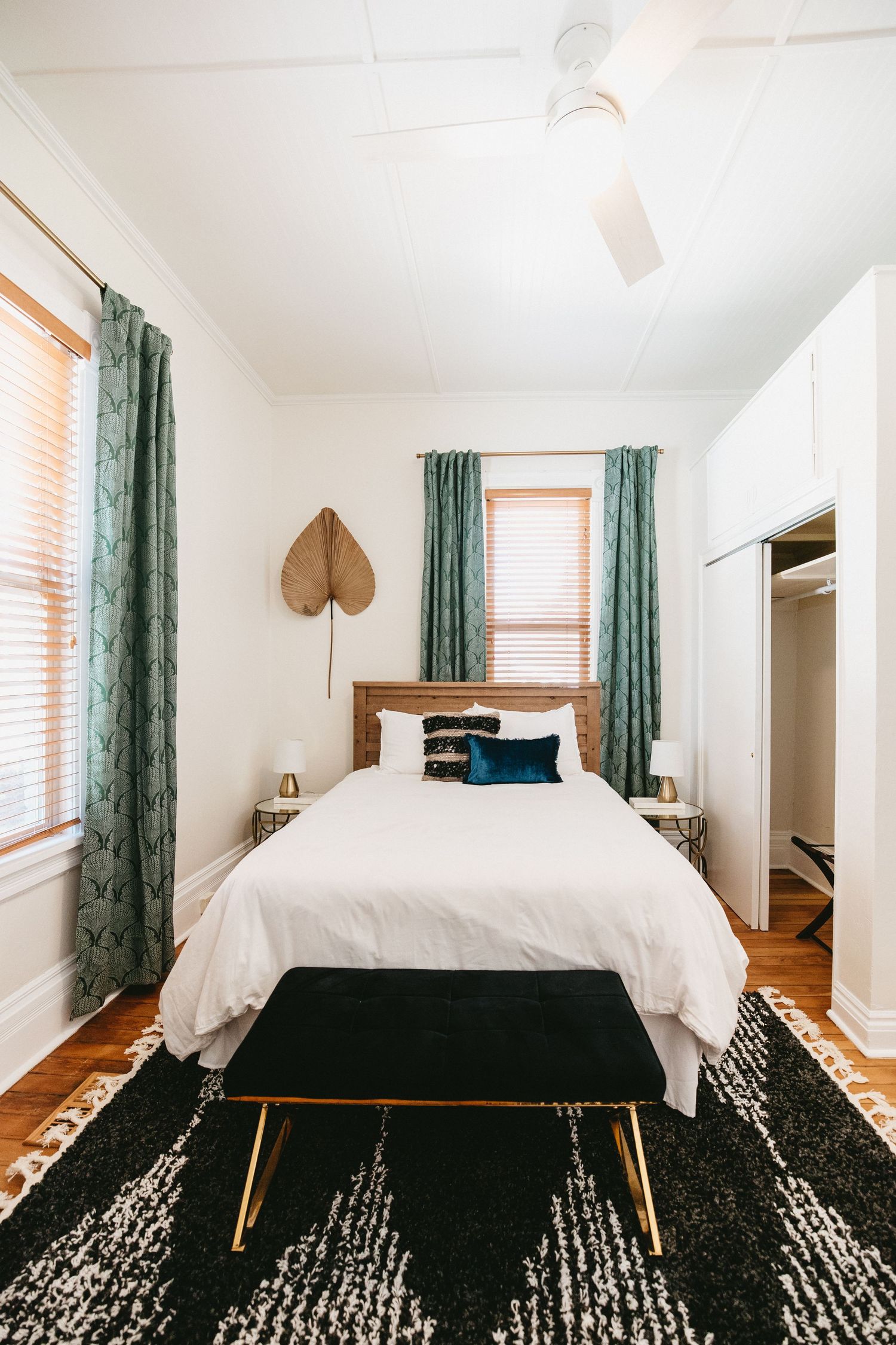 a clean, bright bedroom with fresh white linens, green drapes, a black and white rug, with minimal and modern touches throughout the room