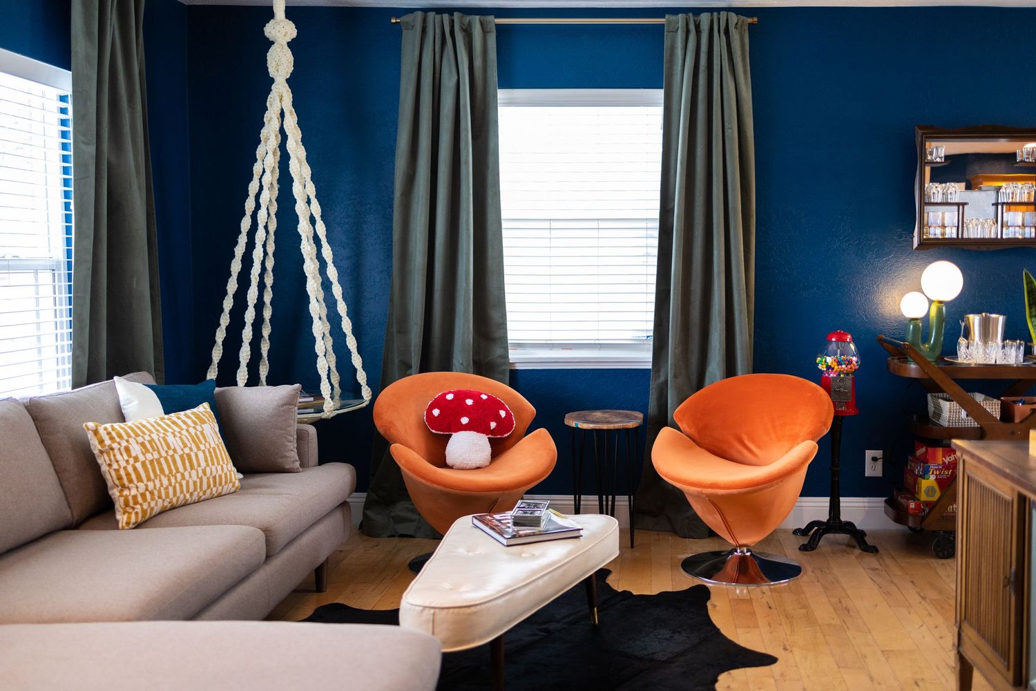 Living room with dark blue walls, a beige L-shaped couch, and two orange swivel chairs, with one mushroom shaped pillow
