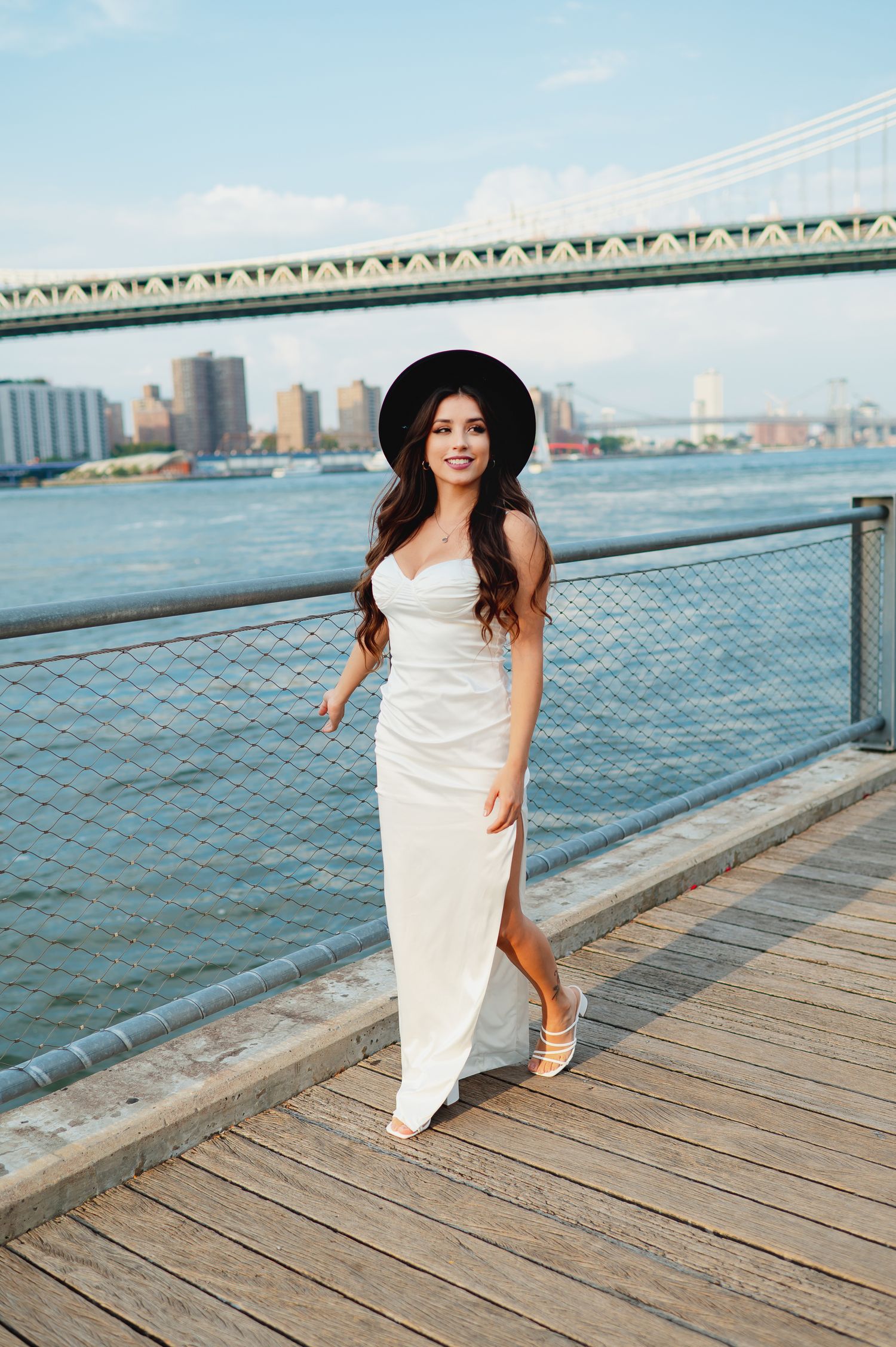 A white dress flows in the breeze on a waterfront boardwalk with a bridge and cityscape backdrop.