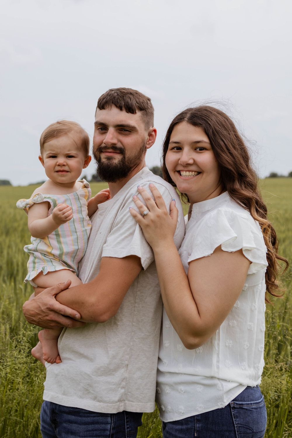 A family in white shirts and jeans embraces while standing in a grassy meadow on a bright summer day.