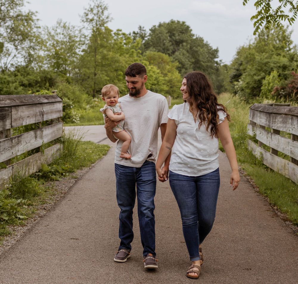 A family walking together holding hands on a wooden bridge pathway through a natural outdoor setting.