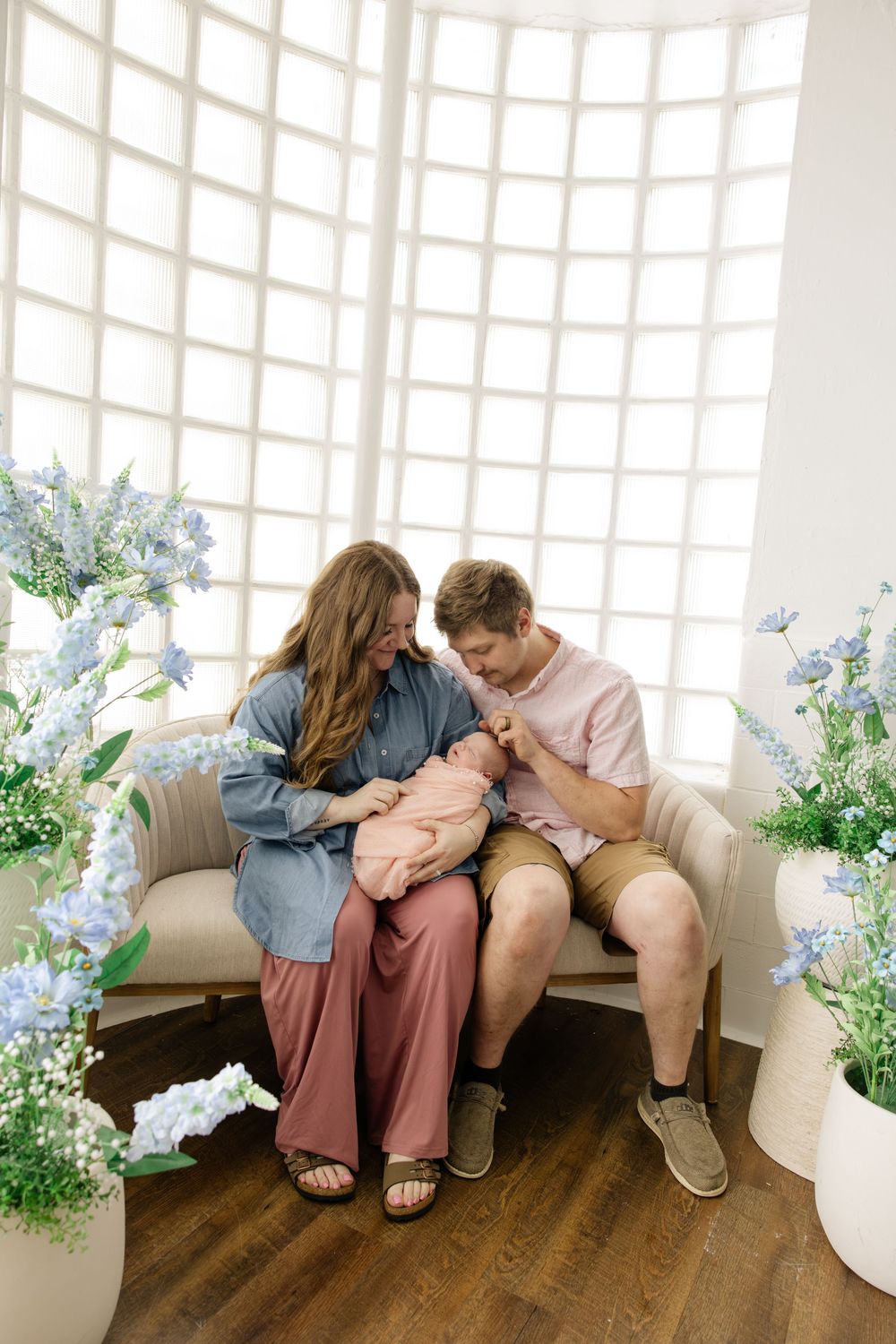 A couple sits on a couch surrounded by blue flowers, lovingly gazing at their newborn baby in a bright studio.