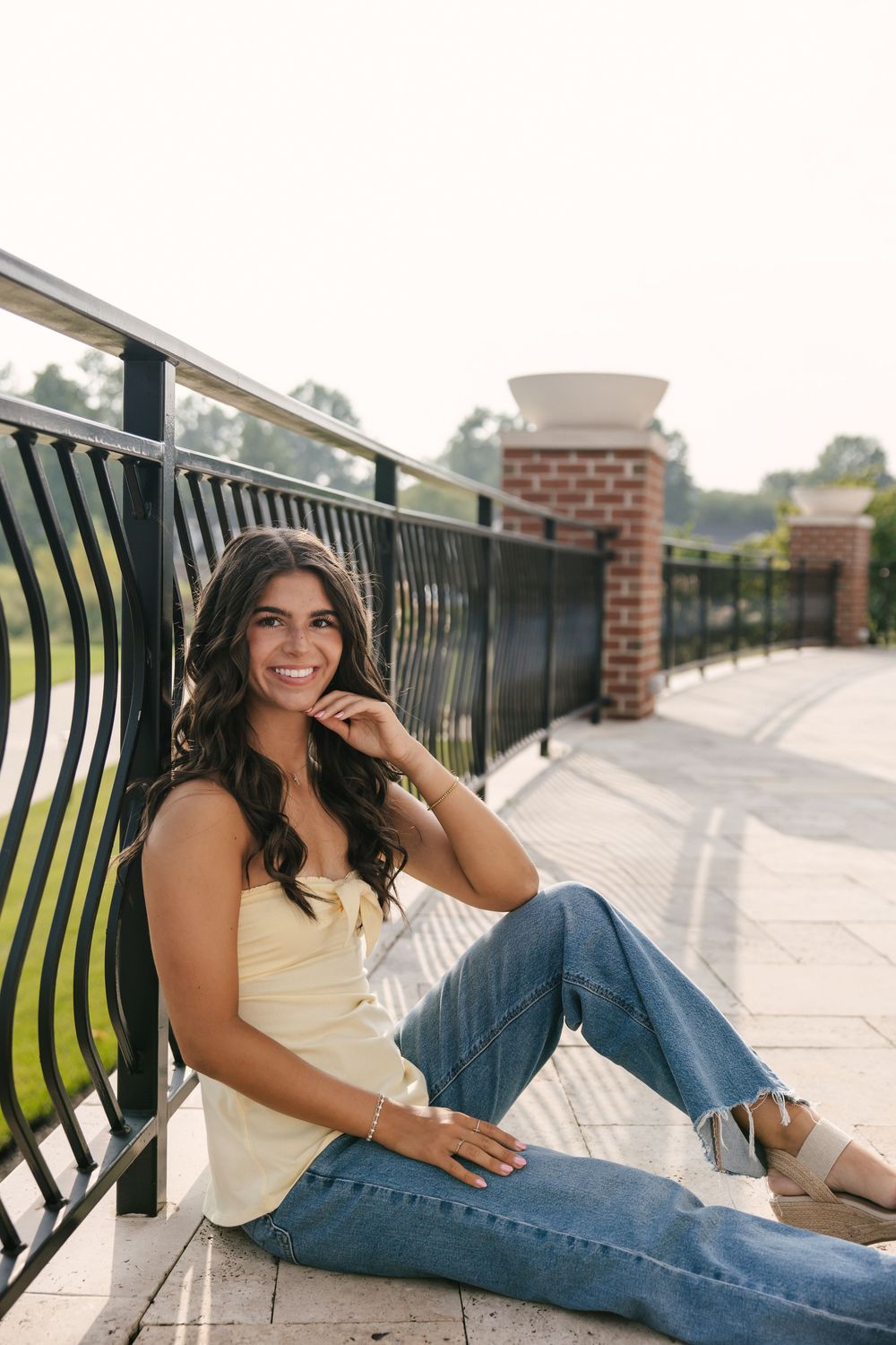 Young woman in yellow top and jeans sits smiling against black iron railing on outdoor brick pathway.