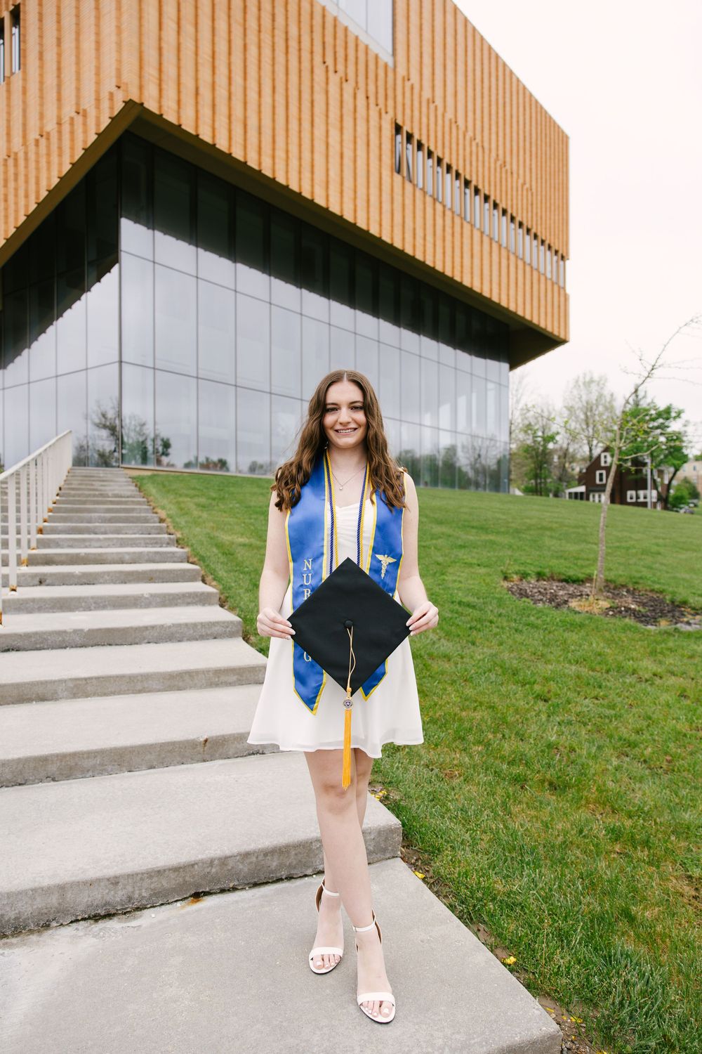 Young woman in white dress holding graduation cap with blue stole, posing on steps outside modern university building.
