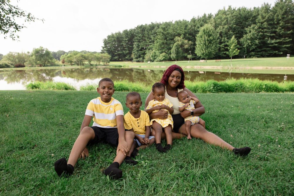 A Black mother sits on green grass with her three young children near a serene pond surrounded by lush trees.