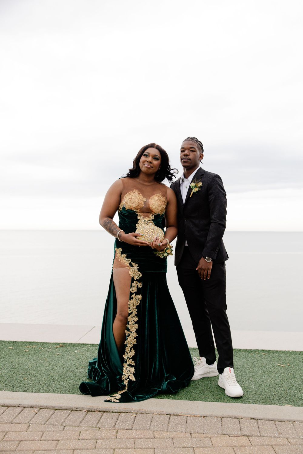 Couple posing outdoors in formal attire, woman in green and gold gown, man in black suit, ocean backdrop.