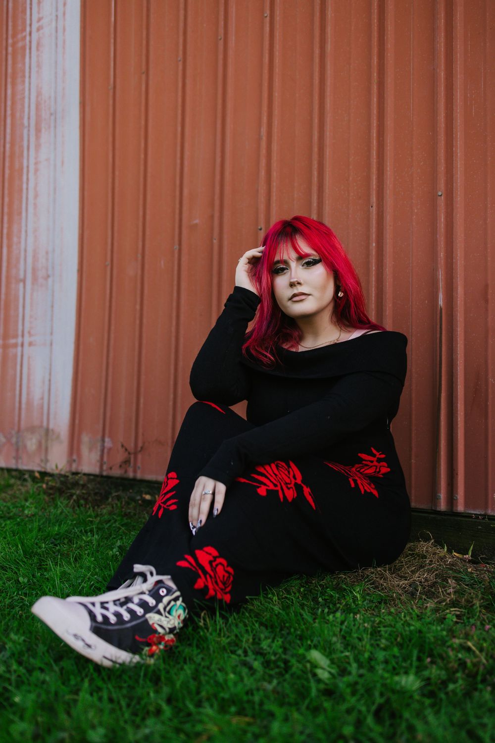 Person with vibrant red hair wearing black clothing lies on grass against an orange metal wall wearing sneakers.