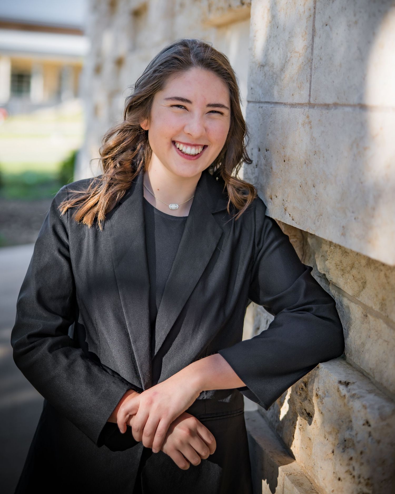 Professional headshot photo showing warm smile wearing black blazer against stone wall background in natural lighting.